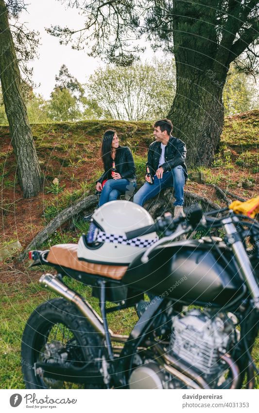 Couple resting from a motorcycle trip attractive couple smiling enjoying vintage alcohol-free beer landscape drinking talking custom built trees retro bike