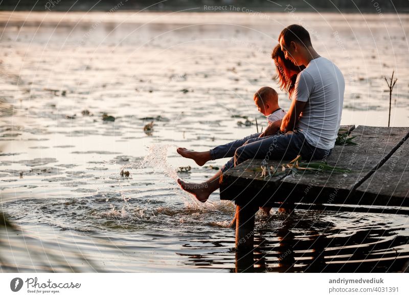 Young parents and their little son sitting on the wooden pier near the lake, at sunset on summer day. family mother young autumn father fall portrait bridge