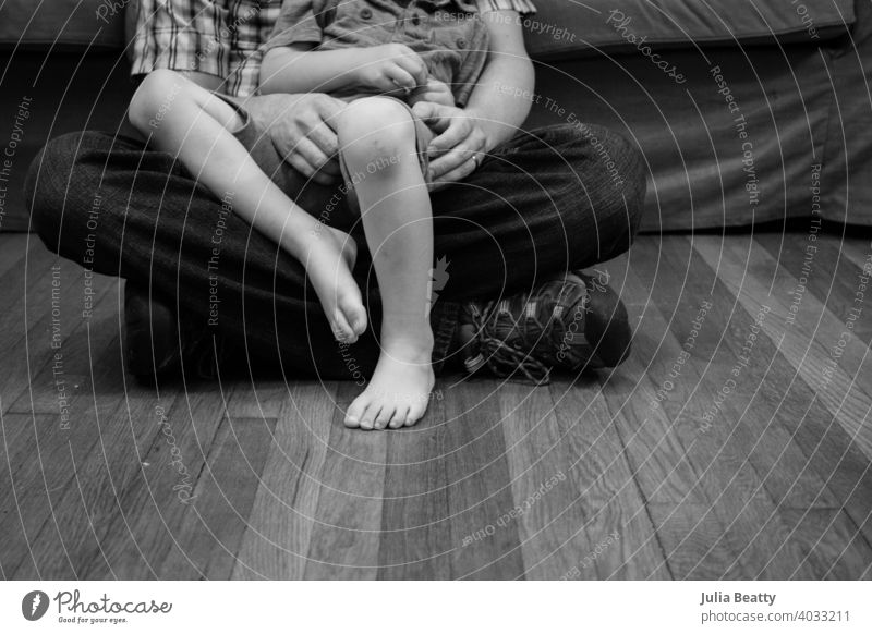 Father and son sitting on hardwood floor; child in dad's lap father childhood single parent bond snuggle boy kid comfort hands feet toes fatherhood floors