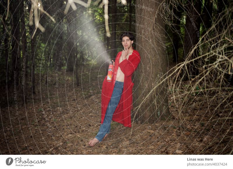 A wild man in the forest is taking down an imaginative fire with a fire extinguisher. Dressed in blue jeans and a red bathrobe he is having fun in these woods. Smoking a cigarette and just enjoying nature.