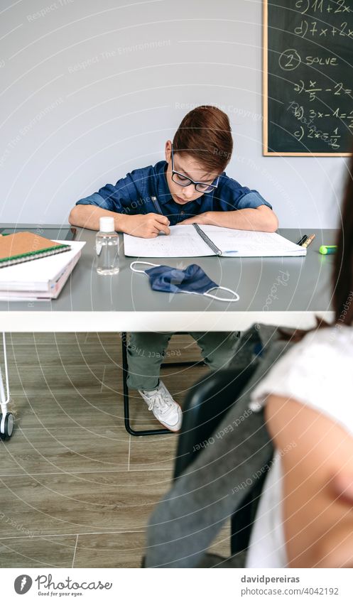 Boy writing in his notebook at school kid student classroom no mask covid-19 safety hand sanitizer virus people epidemic education mask on desk prevention