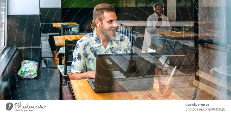 Young man with earphones and laptop in a coffee shop panorama banner happy casual wear looking aside free wifi talking through the glass beard reflection