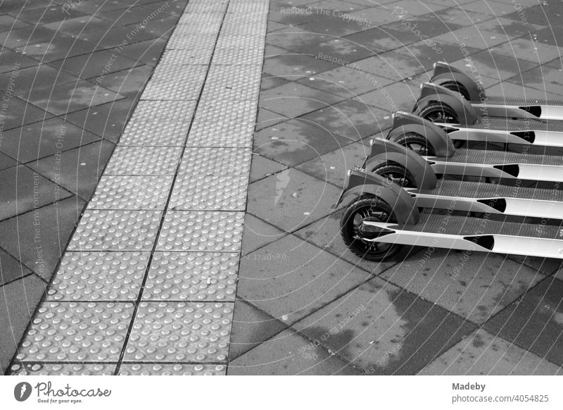 E-scooters lined up for rent next to a tactile guidance system for the visually impaired on grey pavement in the city centre of Frankfurt am Main in Hesse, photographed in neo-realistic black and white