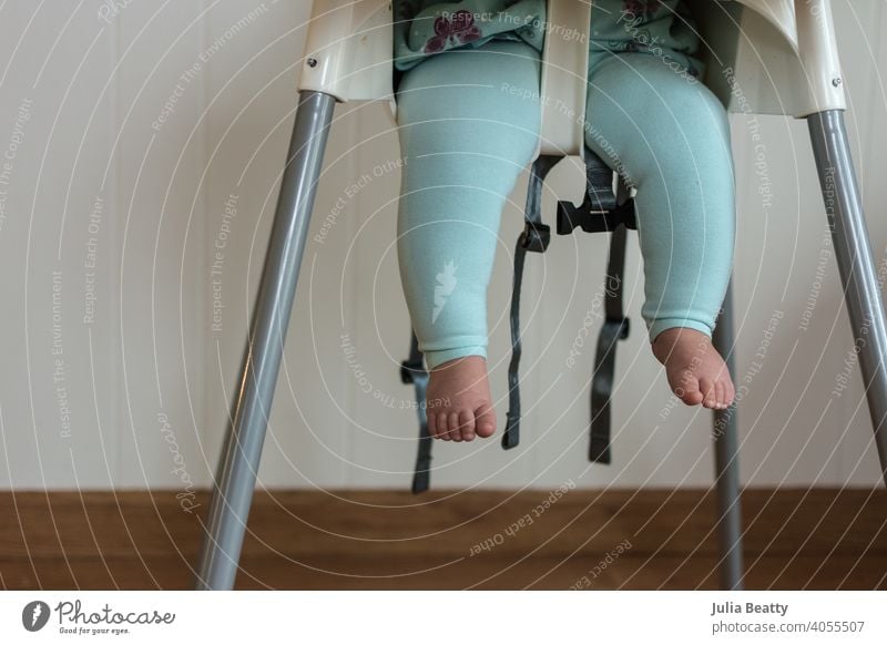 Baby legs dangling from high chair; baby wearing turquoise outfit with bare feet against white wood background toes straps seat belt unbelted fasten unfasten