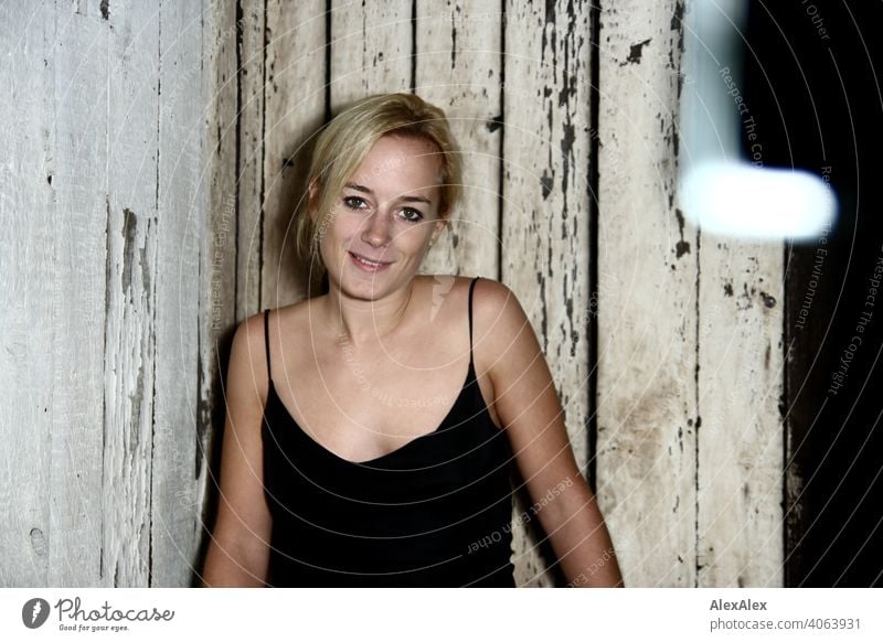 Young woman in black strap dress in front of old board wall in attic Woman Blonde Long-haired pretty Slim Athletic tanned décolleté Face of a woman Smiling