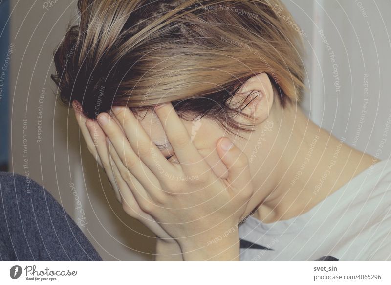 The girl covers her face with her hands. A young girl with blond hair sits in profile on the floor in a room against the backdrop of light-colored furniture and covers her face with her hands.