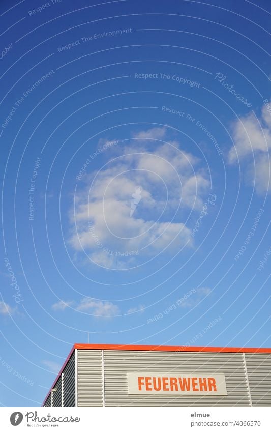 Purpose-built building with grey corrugated iron cladding and a sign with a red inscription "FEUERWEHR" in front of a blue sky with fair weather clouds / 112 / rescue service