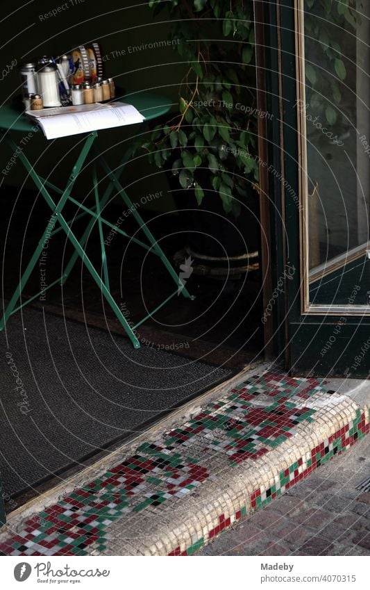 Colorful mosaic on the step at the open entrance to a café with a bistro table with a sugar shaker in the semi-darkness at the Großer Hirschgraben in Frankfurt am Main in Hesse, Germany