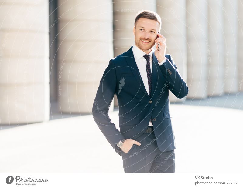 Portrait of successful male manager satisfied with mobile tariffs, makes phone call, uses banking service operator, wears formal suit, stands in urban setting outsie. Professional man worker