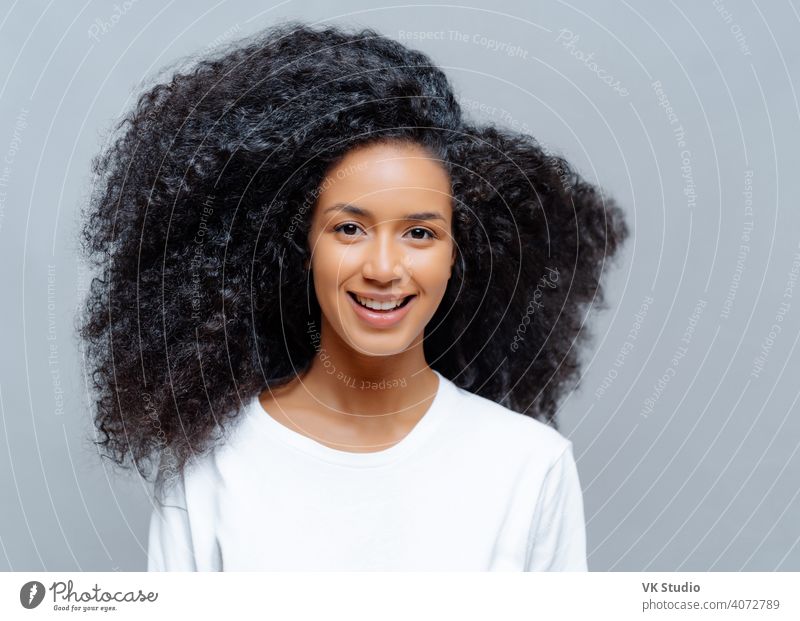 Positive curly woman with natural beauty, dressed in white casual t shirt, has happy expression, looks directly at camera, poses against grey background. Cheerful teenage girl expresses good emotions