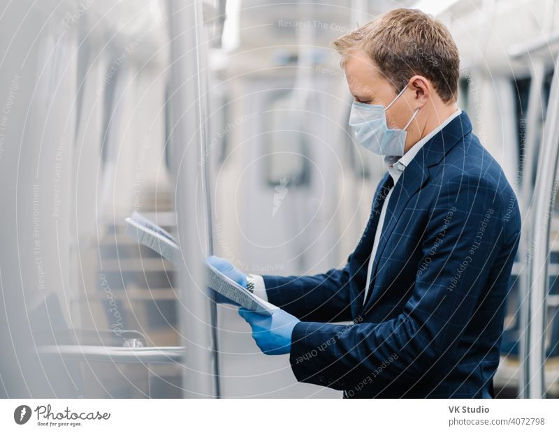 Sideways shot of young elegant man reads newspaper attentively, poses in metro carriage, wears medical mask and disposable rubber gloves from coronavirus. Social distancing, quarantine measures