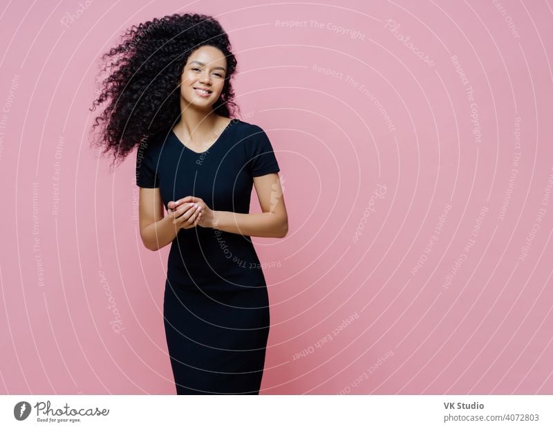 Photo of happy cheerful dark skinned curly woman dresses for casual event, smiles positively, enjoys pleasant talk with man, keeps hands together, stands indoor, free space aside for your text