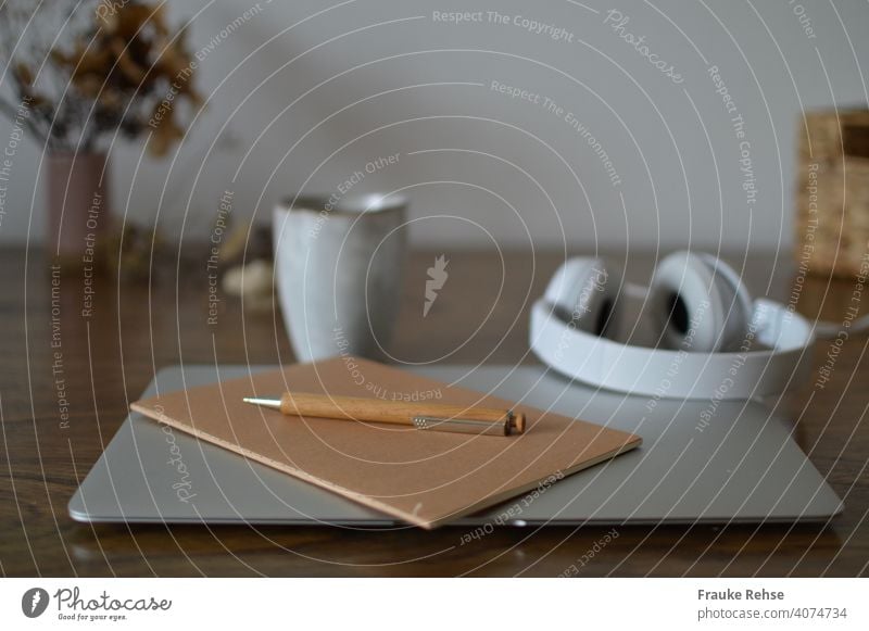 Desk with laptop, notebook, pen in foreground - headphones and mug, tray and flowers in background Home Office Notebook Workplace home office at home thoughts