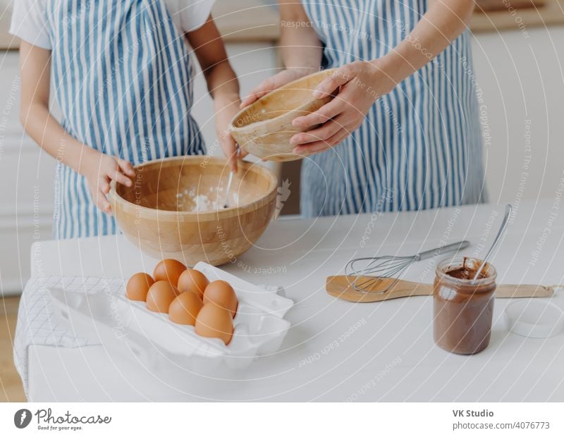 Cropped image of mothers and daughters hands mixing ingredients to prepare dough and bake tasty pastry, stand near kitchen table with eggs, melted chocolate in glass, wear striped blue aprons