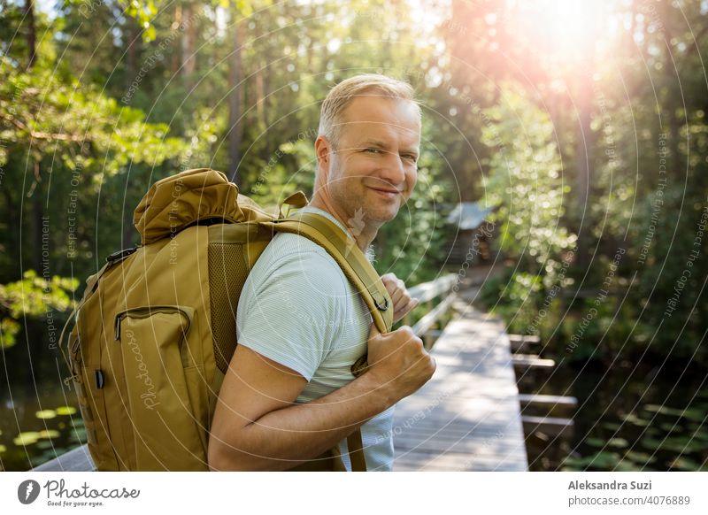 Mature man exploring Finnish nature in summer, walking across the bridge. Hiker with big backpack traveling in forests. Summer Scandinavian landscape of lakes and woods.