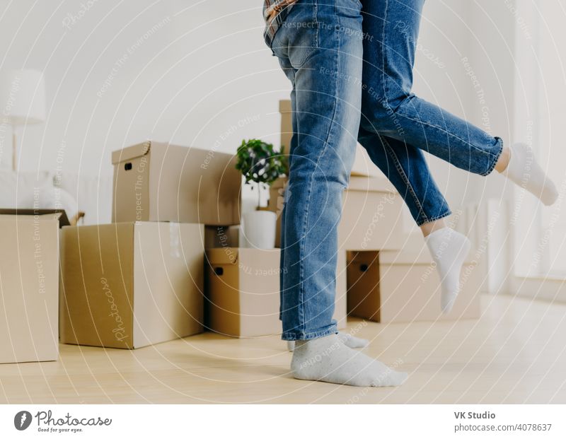 Faceless young couple move in new home, man lifts woman, have fun, surrounded with unpacked cardboard boxes, start new life in recently bought abode. Family, relocation and moving day concept