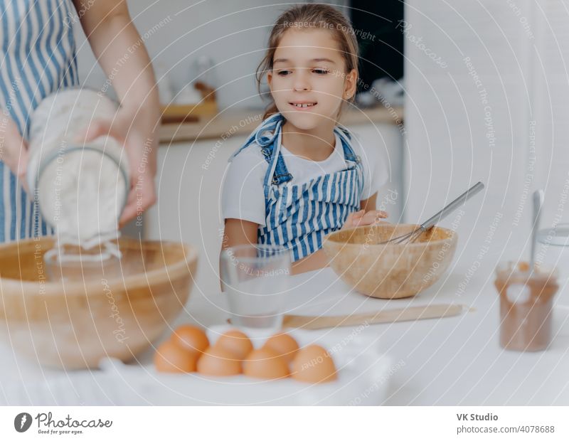 Curious little girl looks how mom prepares dough for pastry, learns to cook, gets culinary experience, wears apron. Faceless woman adds flour in bowl with other ingredients, pose in kitchen with child