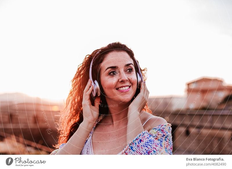 portrait outdoors of a young beautiful woman at sunset listening music on headset and smiling. Lifestyle and music concept casual fun young adult app