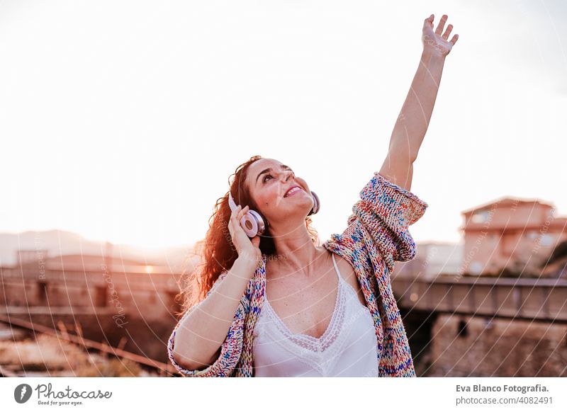 portrait outdoors of a young beautiful woman at sunset listening music on headset and smiling. Lifestyle and music concept casual fun young adult app