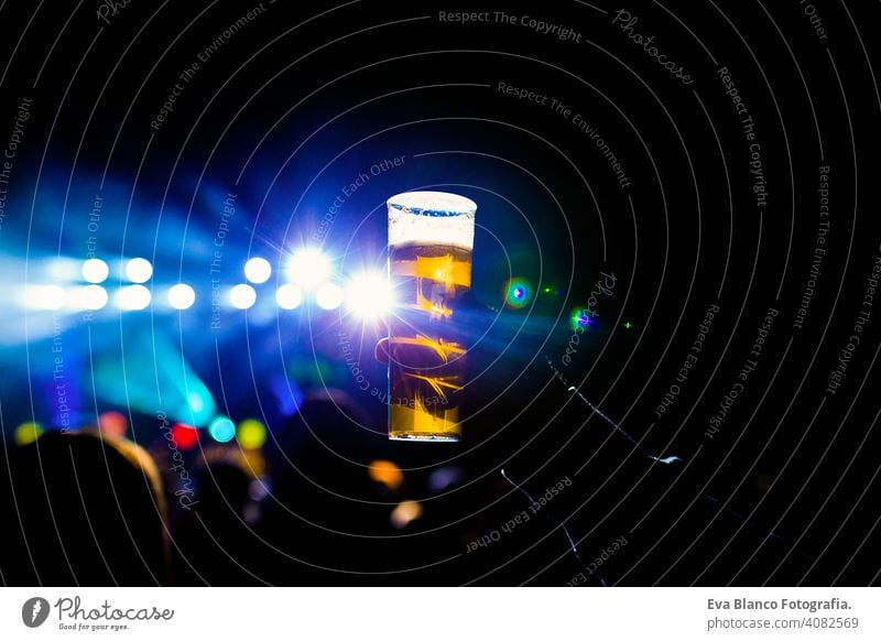 man holding glass of beer in a night concert. Unrecognizable crowd background. blue lights fan people live performer happy music mobile phone dance young