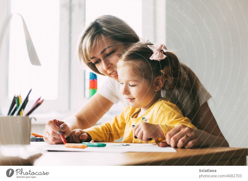 A young mother teaches her little daughter to draw with colored pencils. Time together, creativity, education. Side view girl child mom drawing paint table