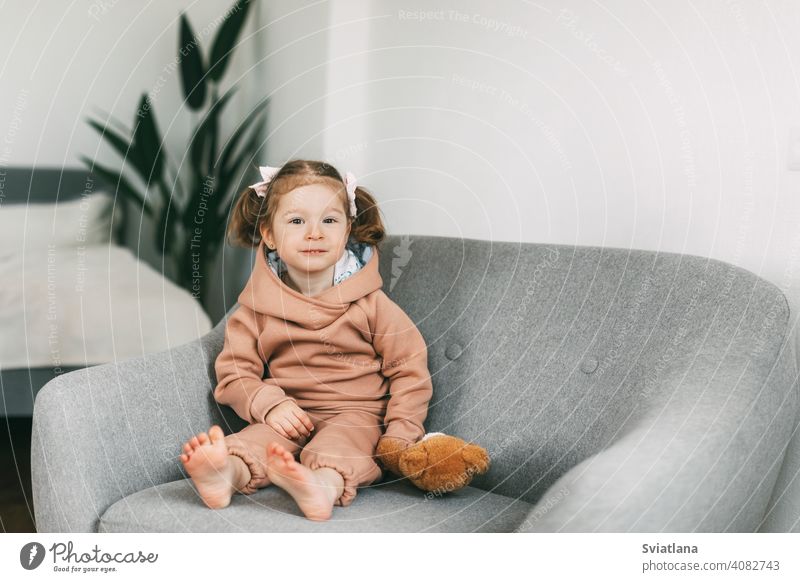 A little charming girl sits in a chair with her toy bear and smiles small kid cute sitting child childhood teddy bear happy fun adorable beautiful young one