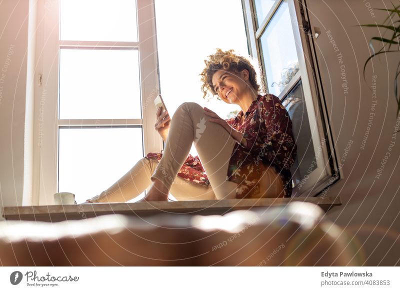 Young woman sitting on window sill and using smartphone happy smiling cheerful apartment leisure bedroom house home alone people caucasian adult indoors person