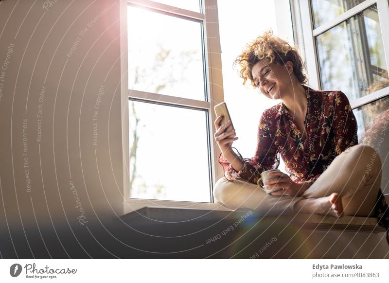 Young woman sitting on window sill and using smartphone happy smiling cheerful apartment leisure bedroom house home alone people caucasian adult indoors person