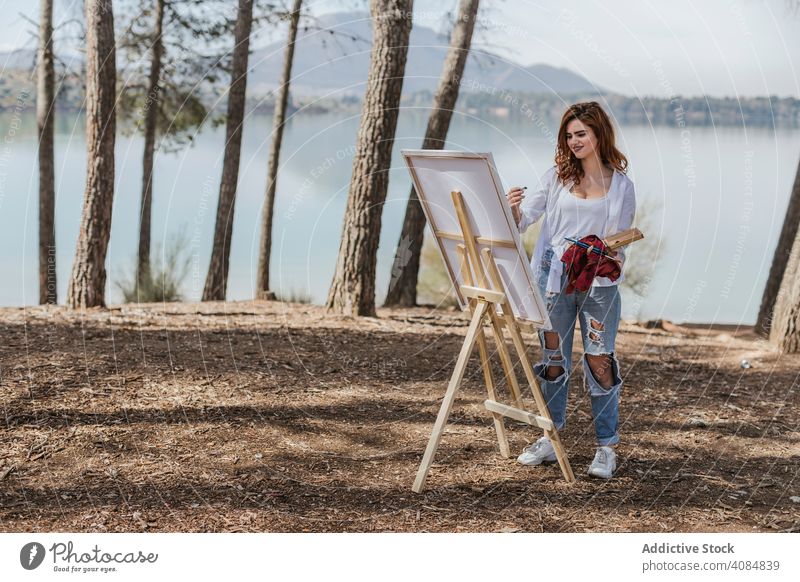 Young woman painting in countryside artist nature lake easel palette landscape summer lifestyle leisure female casual focused concentrated skill talent