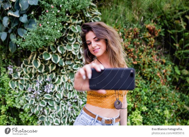 young blonde beautiful woman at the street taking a picture with mobile phone and smiling. Lifestyle outdoors. Summertime, Green background smart urban happy