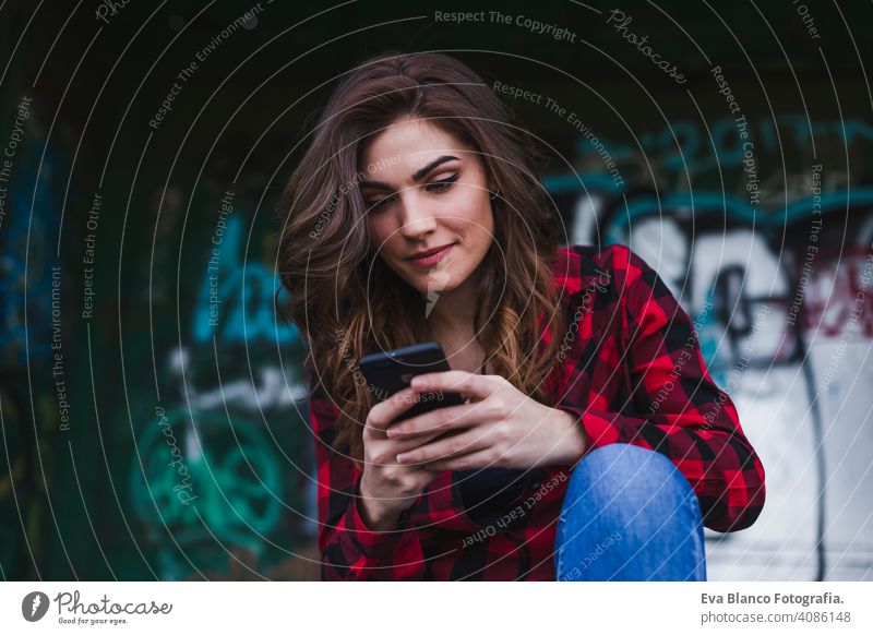 young beautiful woman using mobile phone. Urban graffiti background. Outdoors lifestyle.Technology urban hat standing attractive fence cityscape selfie clouds