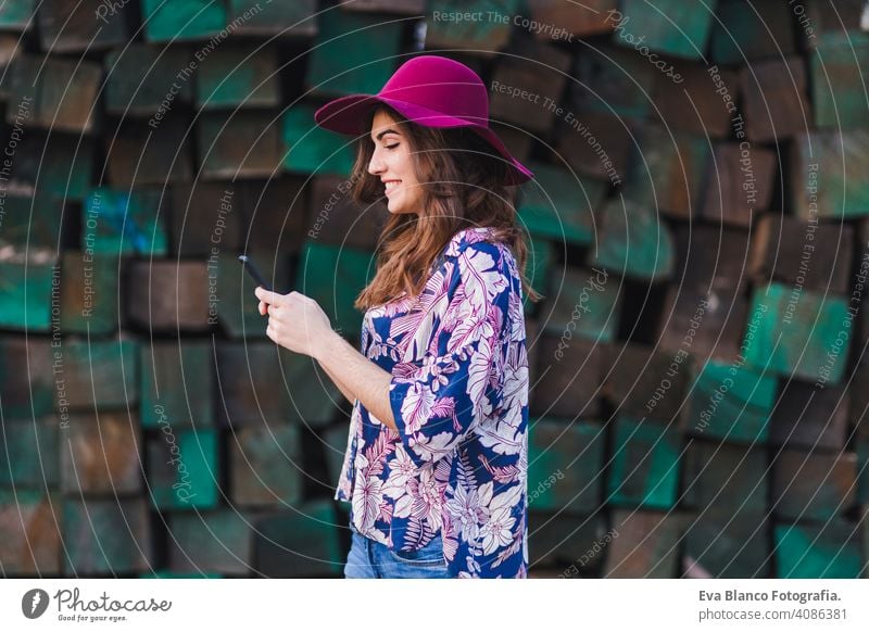 portrait of a young beautiful woman wearing casual clothes and a modern hat and using her mobile phone. She is standing over green wood blocks background and smiling. Outdoors lifestyle.