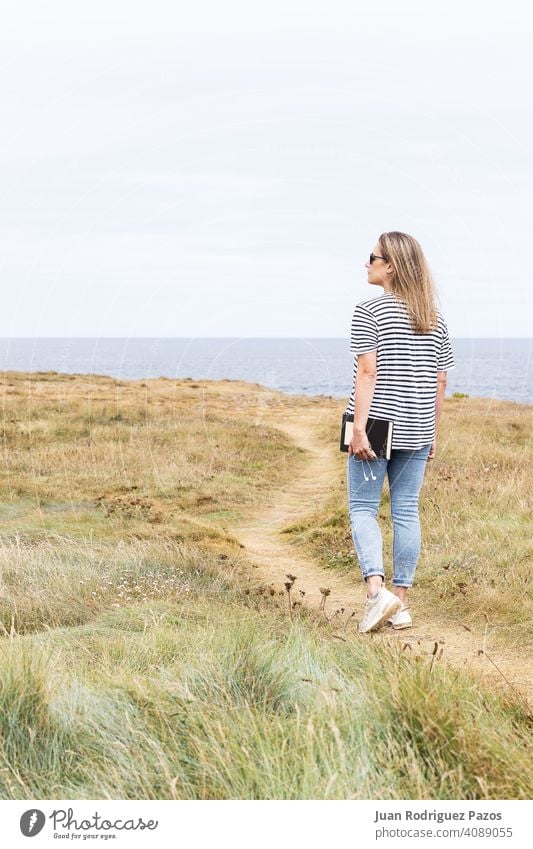 Woman walking along a path in front of the sea holding a book and headphones in her hand read outside relax relaxed enjoy literature mature novel weekend