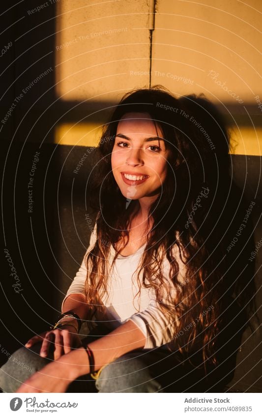 Smiling woman sitting in spot of light seating portrait looking at camera sunlight shadows smiling young casual city street female wall sensual exterior town