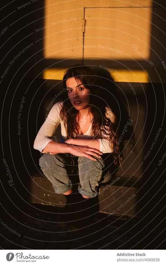 Smiling woman sitting in spot of light seating portrait looking at camera sunlight shadows smiling young casual city street female wall sensual exterior town