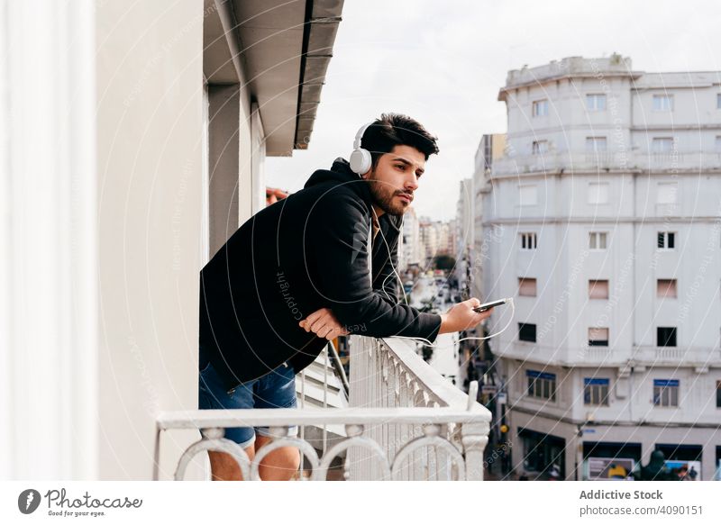 Young happy man holding smartphone on balcony headphones headset leaning railing terrace young male smiling joy cheerful handsome successful cool beard mustache