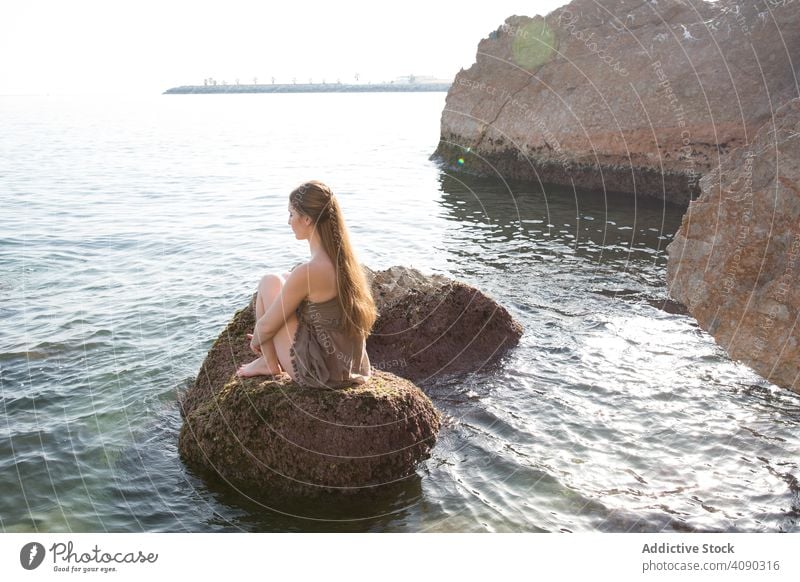 Barefoot woman sitting on rock and looking at sea ocean barefoot young fabric wind water boulder female vacation travel trip tourism sensual elegant dress cloth