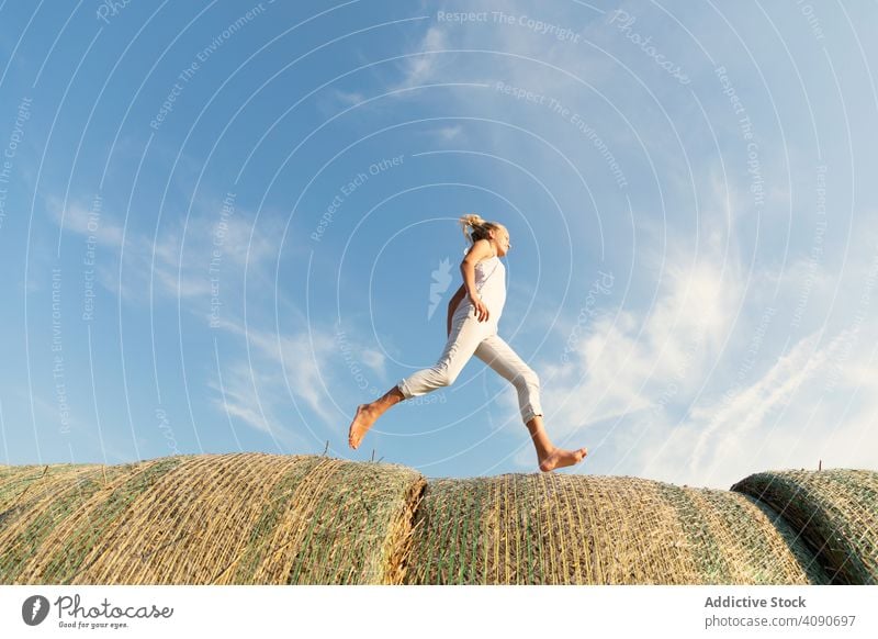 Barefoot girl running on haystacks farm sky clouds sunny daytime nature lifestyle leisure teen kid child barefoot dried grass straw rolls countryside rustic