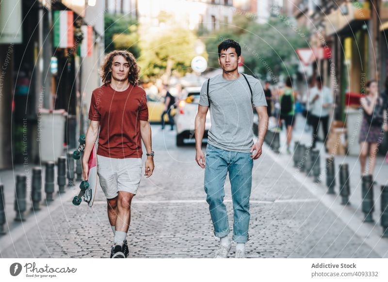 Cool multiracial male friends chatting while walking on street with longboard casual road relaxed interested carefree confident diverse men positive gesturing