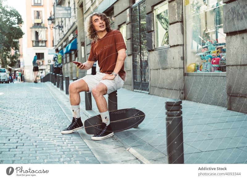 Amused man with longboard talking on phone while resting on street amused casual laugh gesture cheerful using speak smartphone sunny baluster pavement sit