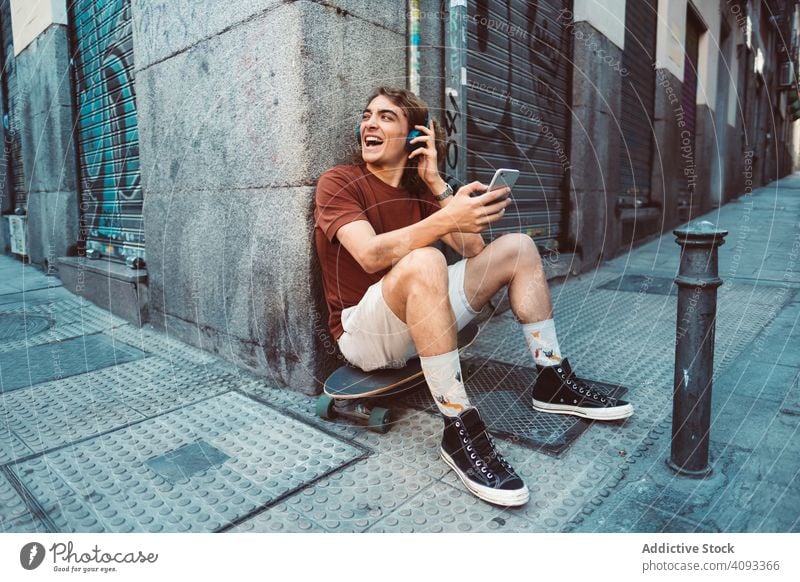 Relaxed man in headphones using smartphone while resting on longboard on street relaxed carefree generation millennial casual browsing sit building city urban