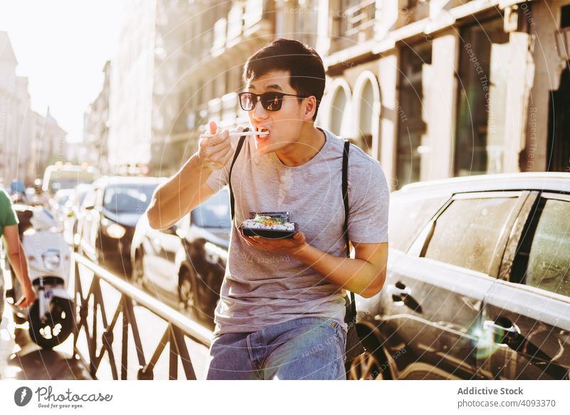 Man eating takeaway food on city street man chopsticks street food hungry asian food urban sunny food truck enjoying ethnic fast food sunglasses young adult