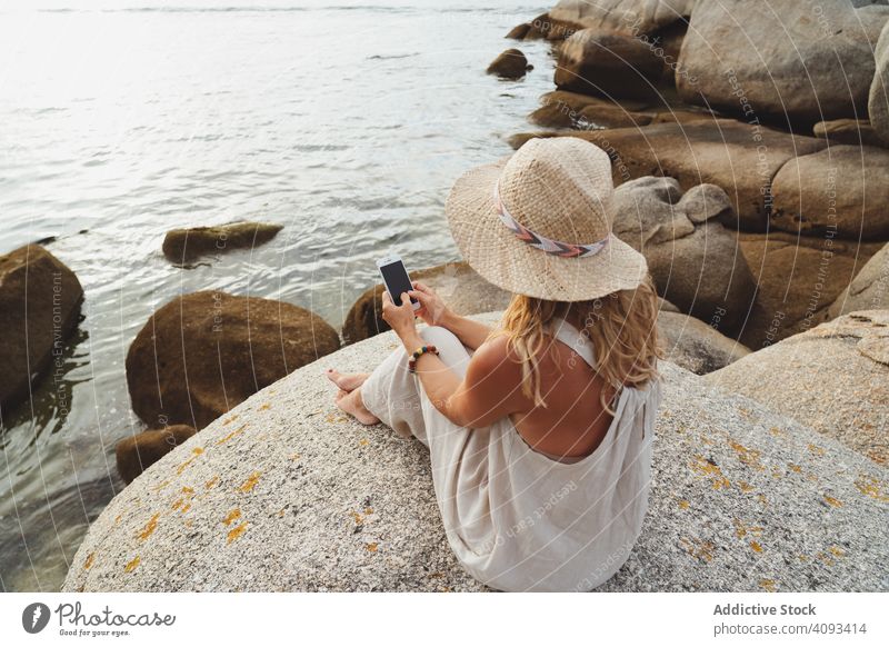 Contemporary woman with phone chilling on seashore smartphone coast alone enjoy escape travel seaside solitude remote wireless straw hat gadget trip vacation