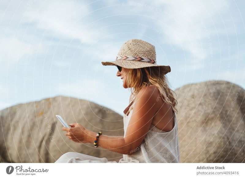 Contemporary woman with phone chilling on seashore smartphone coast alone enjoy escape travel seaside solitude remote wireless straw hat gadget trip vacation