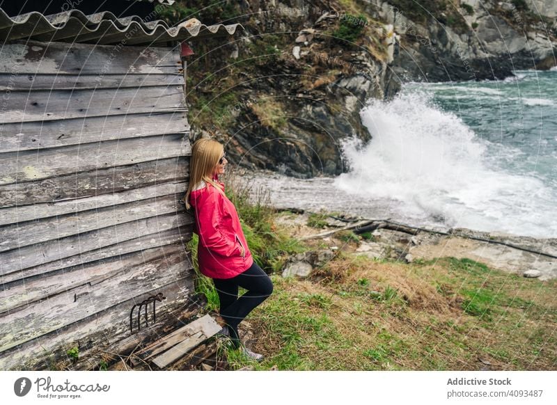 Woman in red raincoat leaning on house at shore looking at water woman seaside wave vacation travel old harbor alone building architecture relaxation thinking