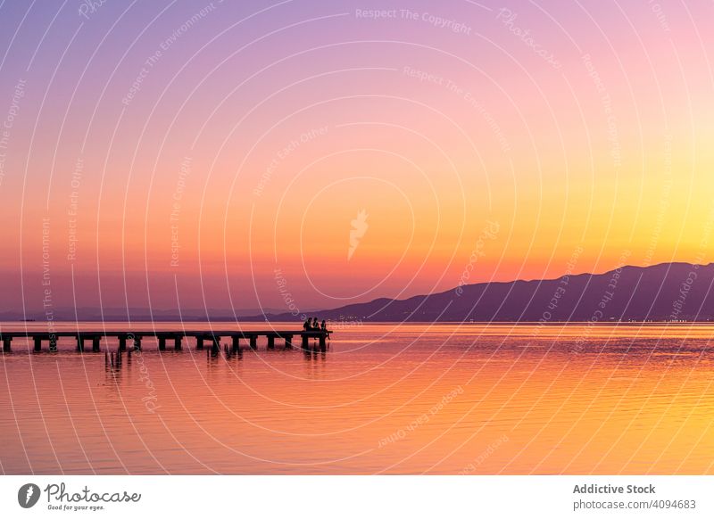 Anonymous tourists sitting on pier during sundown lake sunset traveler silhouette tranquil cloudless sky evening bright water tourism trip weekend holiday