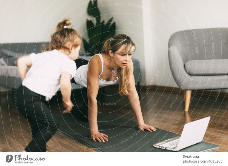 A young mom is doing sports at home and watching a video fitness lesson on a laptop, and her little baby is sitting on a chair. Home training, fitness, sports