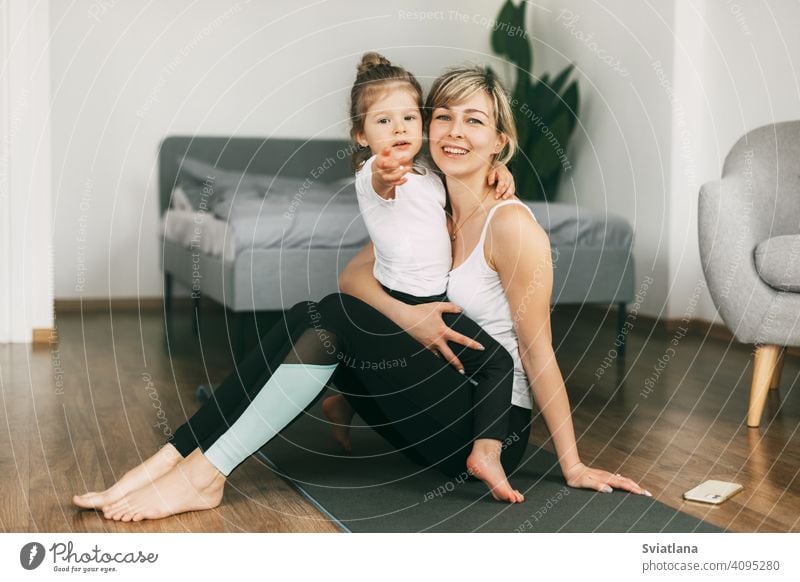 A little girl is sitting on her mother's lap, resting after a fitness exercise, hugging her, smiling, looking at the camera. The concept of a healthy family lifestyle