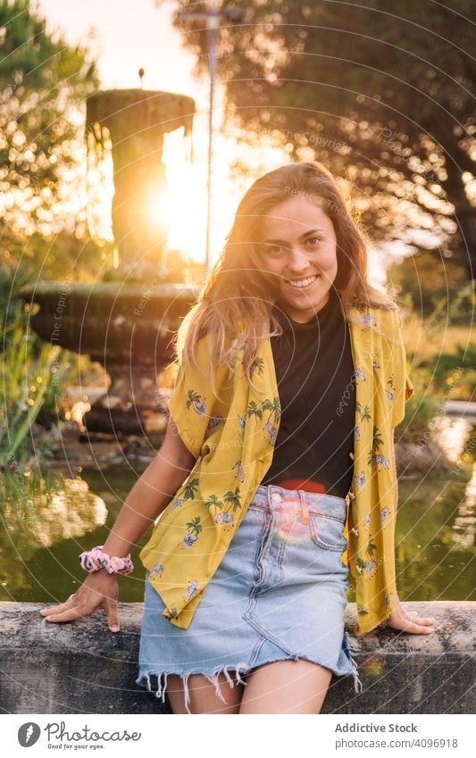 Joyful female relaxing in park woman sunset fountain harmony charming serene smile lean casual wear summer happy holiday leisure nature resort playful rest