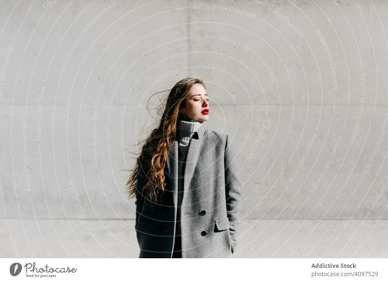 Young woman in coat on windy day stylish street closed eyes wall city building female urban fashion cool young model outfit warm weather exterior lady long hair